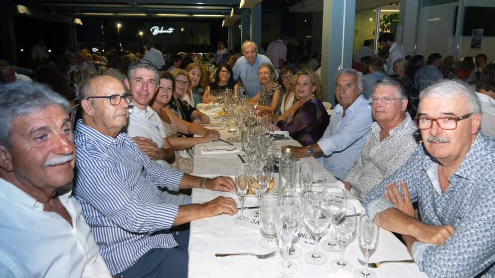 Grupo de personas, en su mayoría adultos mayores, sentados en una mesa larga en un restaurante mientras disfrutan de una cena. La mesa tiene varias copas y vajilla, y la escena es en la noche con mucha iluminación y otros comensales en el fondo.