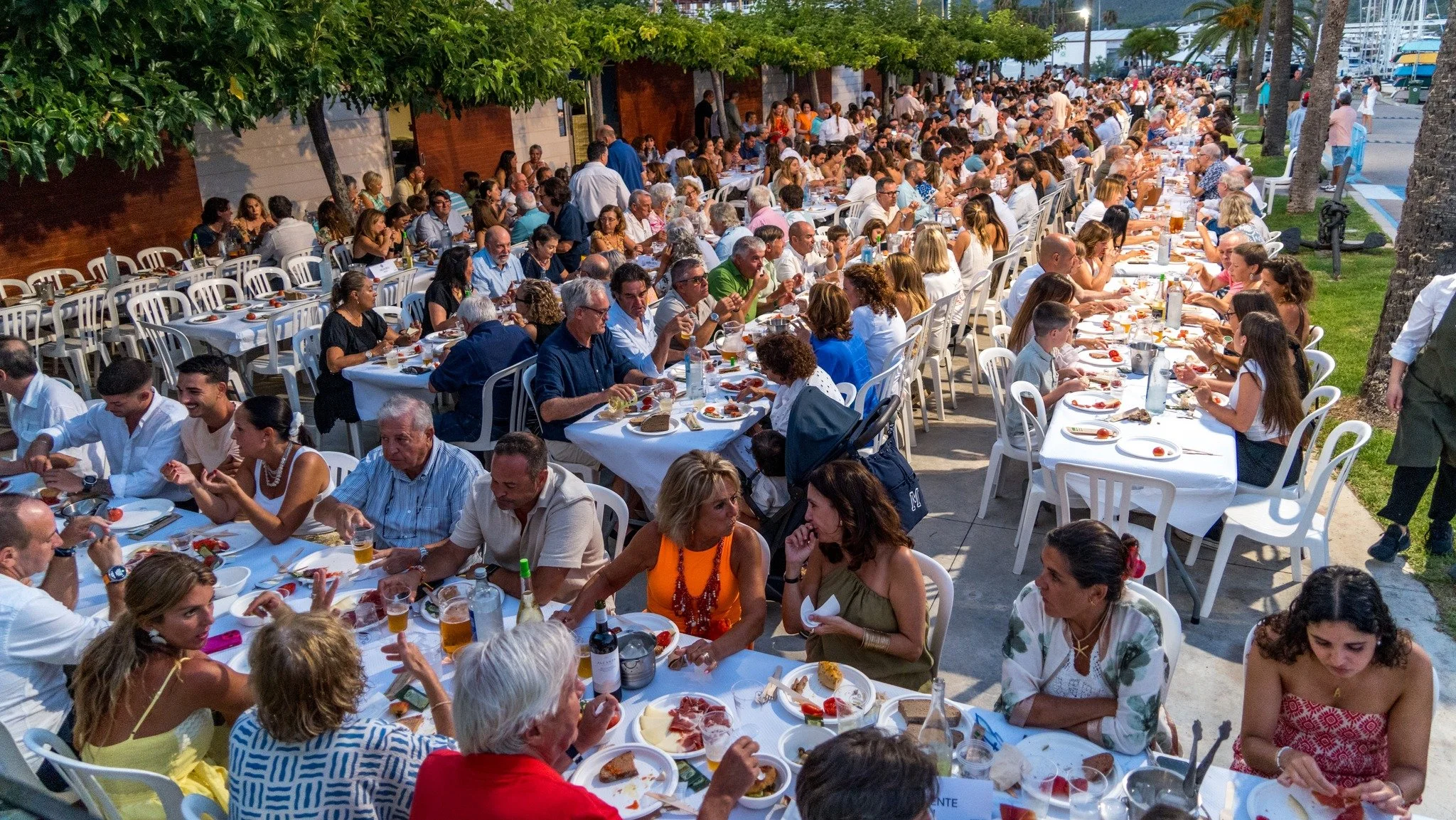 Gran reunión al aire libre con muchas personas sentadas a mesas largas, disfrutando de comida y bebida, en un evento social nocturno en un espacio abierto con árboles y palmeras.