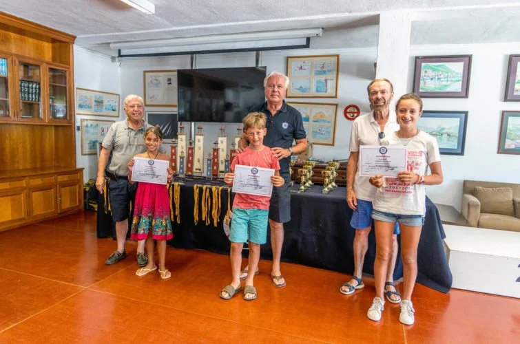 Cuatro niños y dos adultos posando con certificados y trofeos en un salón, celebrando un logro o evento.