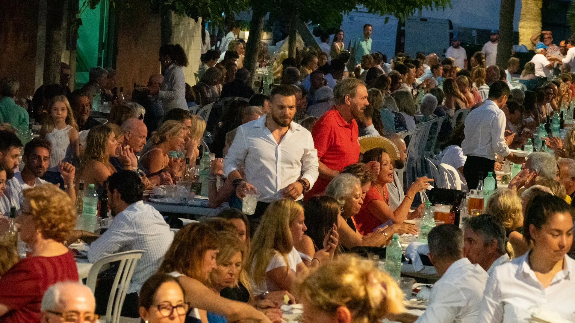 Muchísima gente sentada en mesas al aire libre en una fiesta o celebración por la noche, con árboles y algunas paredes visibles.