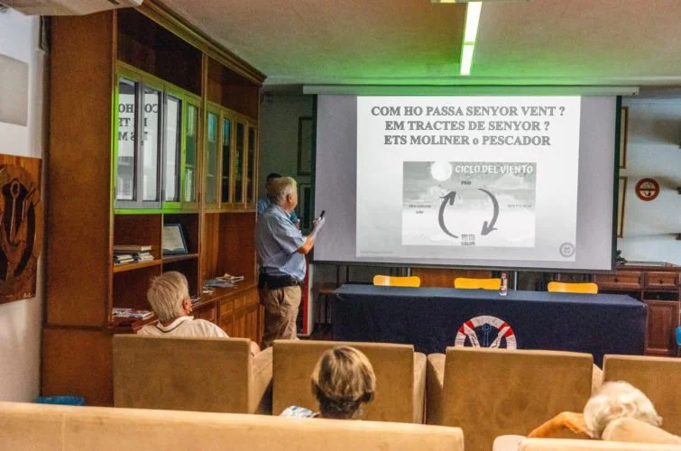 Una presentación en una sala con varias personas sentadas, un hombre de cabello gris y camisa de cuadros dando la charla, un proyector mostrando un texto en español y una imagen de un ciclo del viento.