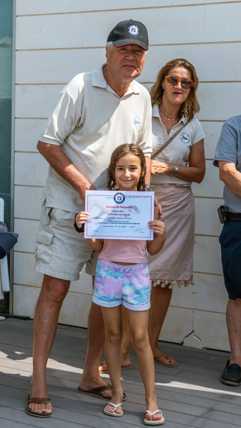 Una niña sonriente con traje de baño de colores pastel sostiene un diploma en un evento de natación, acompañada por un hombre mayor y una mujer, ambos con camisetas blancas con logotipos relacionados con la natación, en un entorno exterior.