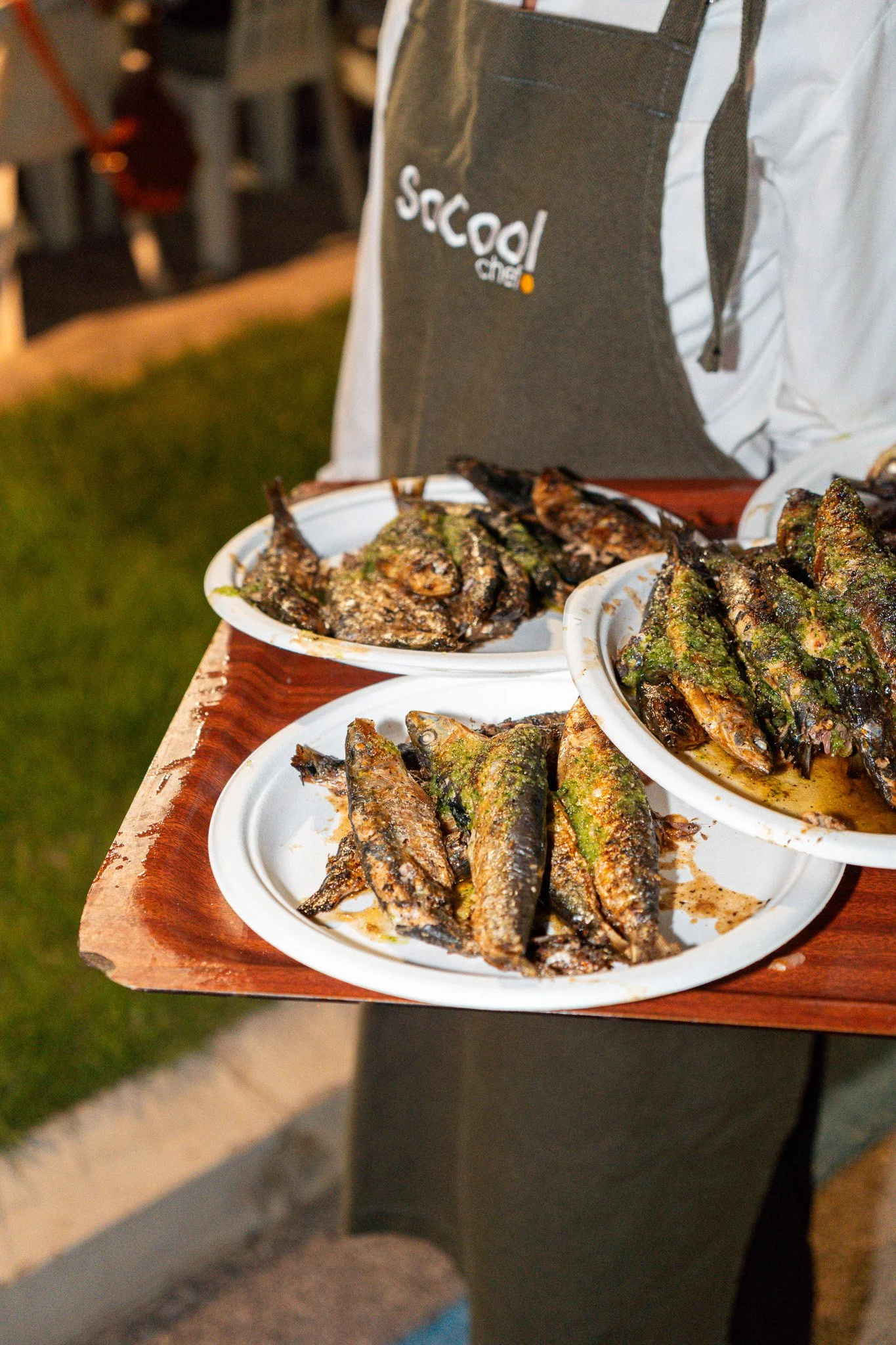 Plato con diferentes especies de pescado asado, algunos con hierbas y especias, servido en platos de papel, siendo llevado por una persona con delantal oscuro y camisa blanca.