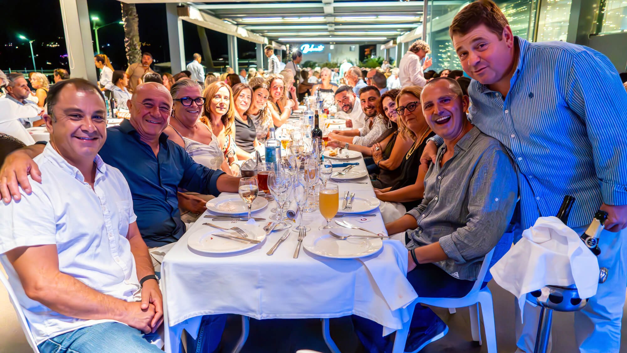 Grupo de personas sentadas en una mesa larga en un restaurante, disfrutando de una cena, algunos sonrientes y otros en conversación, en un ambiente nocturno con iluminación cálida.