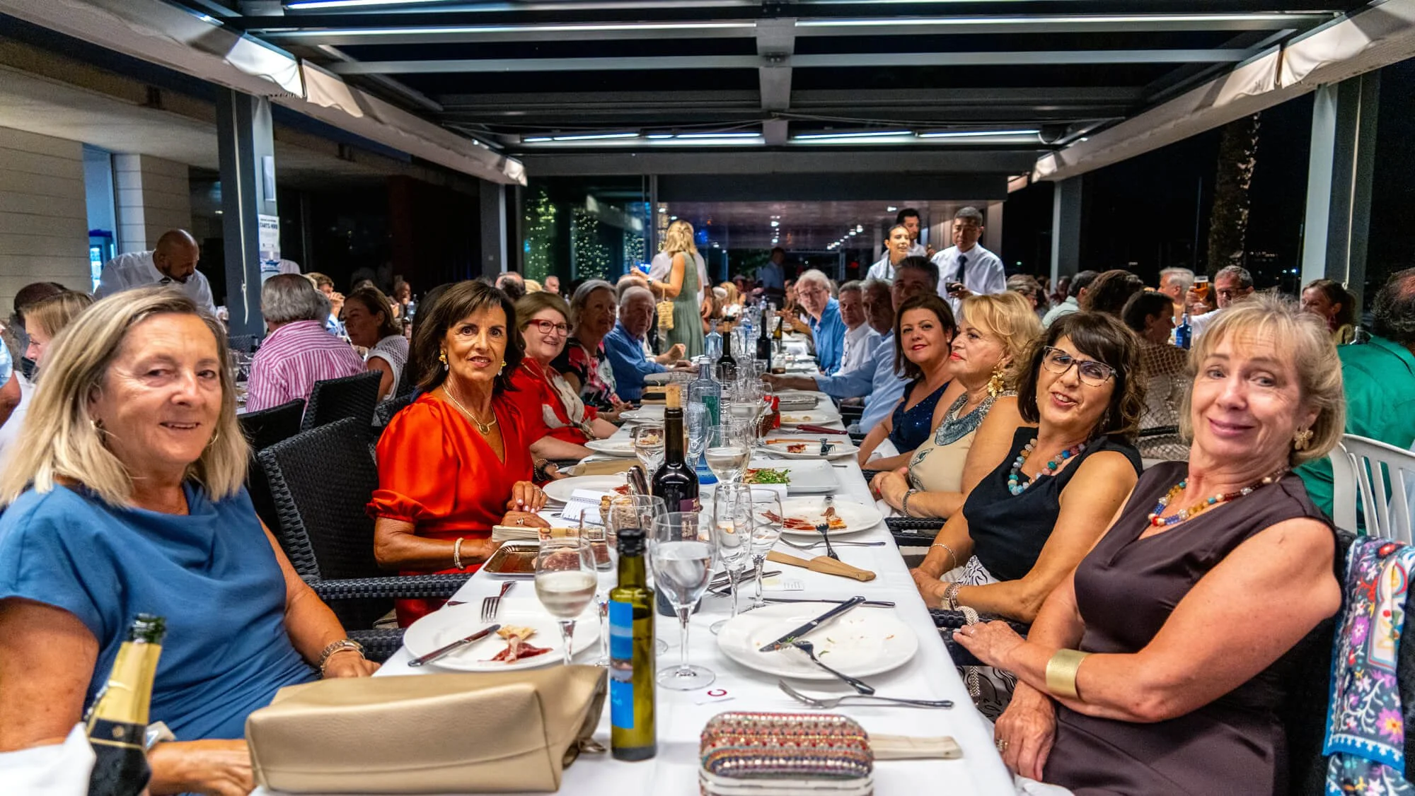 Grupo de personas sentadas en una mesa en un restaurante o evento social, con comida y bebidas sobre la mesa, en un ambiente cubierto durante la noche.
