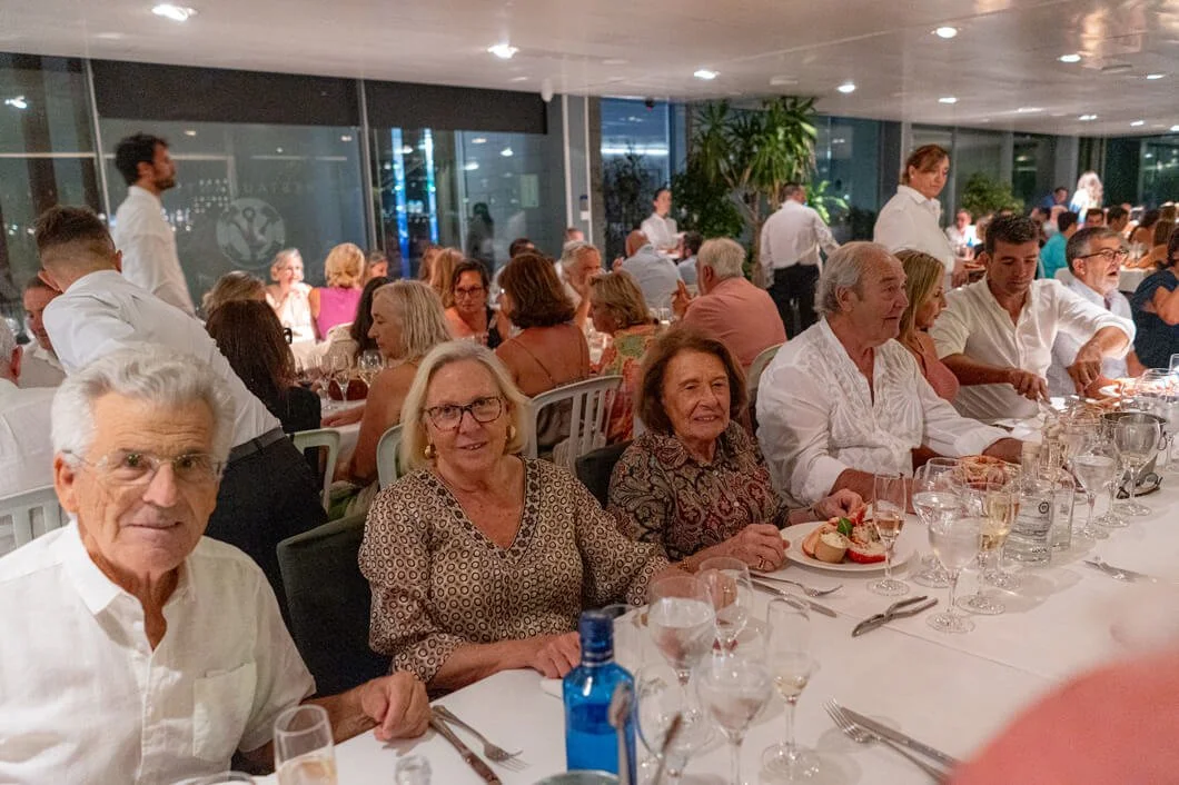  Personas adultas sentadas en una mesa en un restaurante o banquete lleno de gente, con platos y vasos en la mesa, en un ambiente elegante y concurrido.