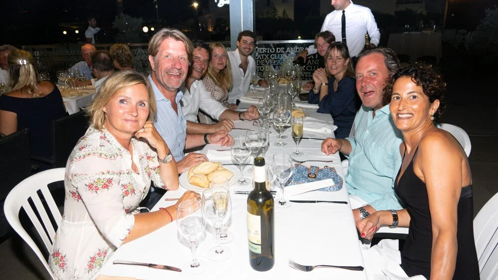 Grupo de personas sonriendo y disfrutando una comida en una mesa larga en un evento social al aire libre por la noche.