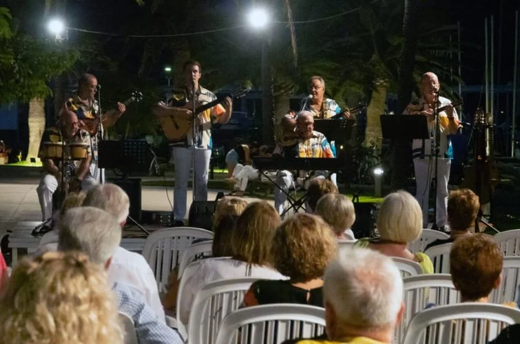 Un grupo de músicos tocando en un escenario al aire libre durante la noche, con un público sentado en sillas plásticas observando el espectáculo.
