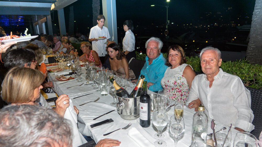 Personas sentadas en una mesa larga en un restaurante, disfrutando de una cena con varios comensales y comida, en un ambiente elegante y social por la noche.