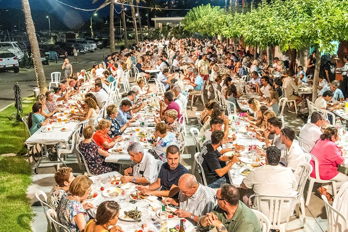 Imagen de una gran comida al aire libre en la noche, con muchas mesas largas y personas sentadas cenando, adornadas con árboles y luces decorativas.