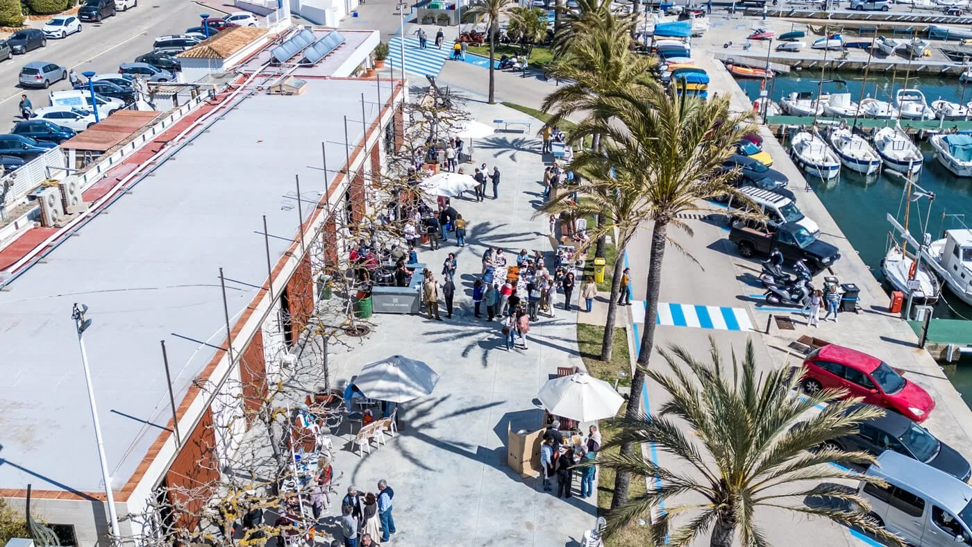 Imagen de una zona de puerto con botes amarrados, autos estacionados y personas en una mesa con sombrillas, en un día soleado con palmeras y cielo despejado.