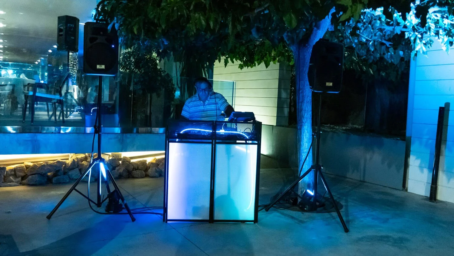 Un DJ tocando en una fiesta nocturna iluminada con luces azules y de colores, con un árbol decorado en el fondo.