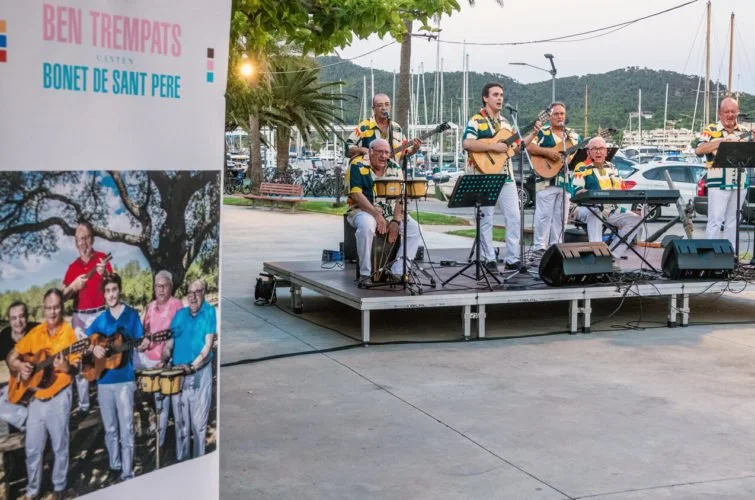 Un grupo de músicos tocando en un escenario al aire libre junto a un puerto con barcos. Un cartel en primer plano promociona a 