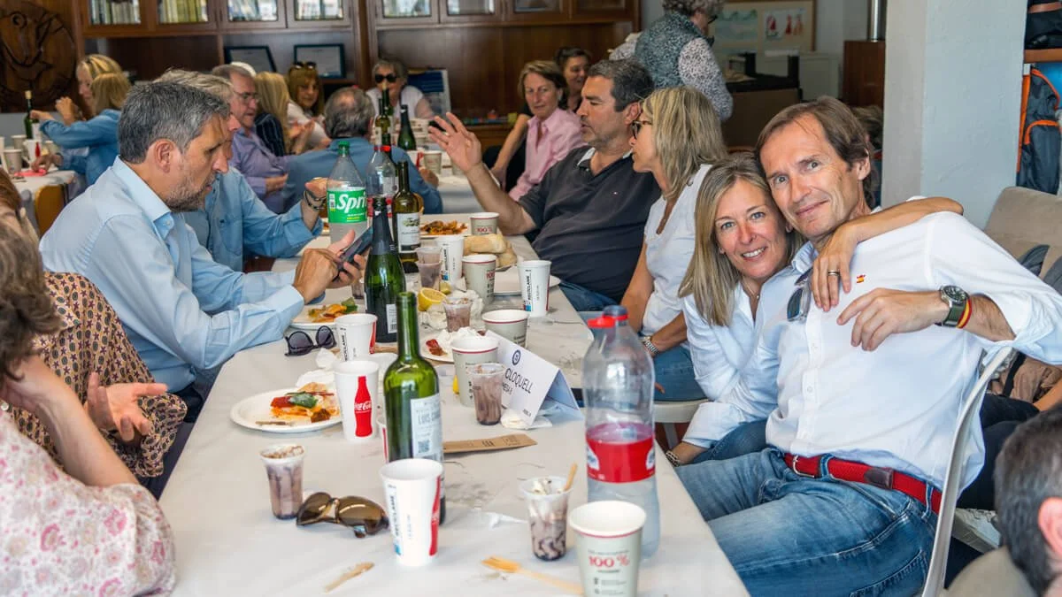 Grupo de personas sentadas en una mesa durante una comida, con varias botellas, vasos y comida en la mesa, en un ambiente interior.