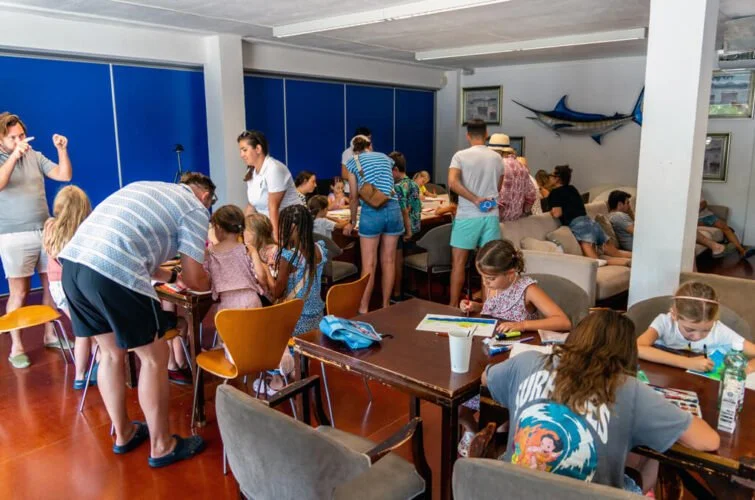 Niños y adultos en una sala de actividades, algunos dibujando y otros conversando, decorada con un pez en la pared.