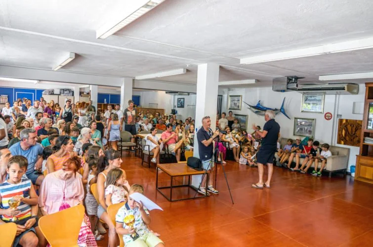 Reunión con muchas personas, incluyendo niños y adultos, en una sala con paredes blancas, decorada con fotos y objetos marinos, donde hay una persona con micrófono y otra haciendo una presentación.