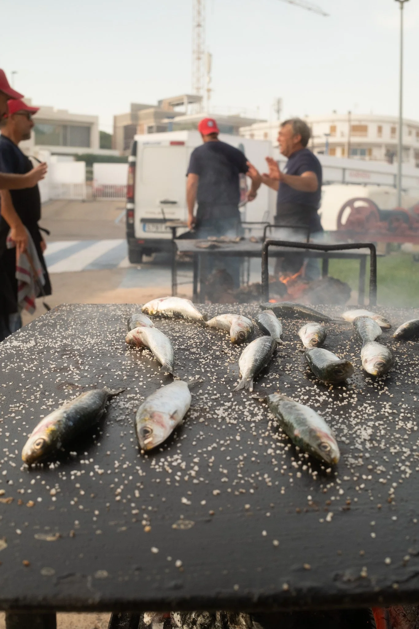 Pescados sobre una superficie, con semillas de sésamo, en primer plano. En el fondo, una parrilla con pescados asándose y personas conversando en un ambiente al aire libre.