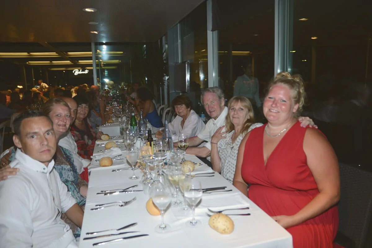 Grupo de personas sentadas en una mesa larga en un restaurante elegante, con comida y copas de vino, durante la noche.