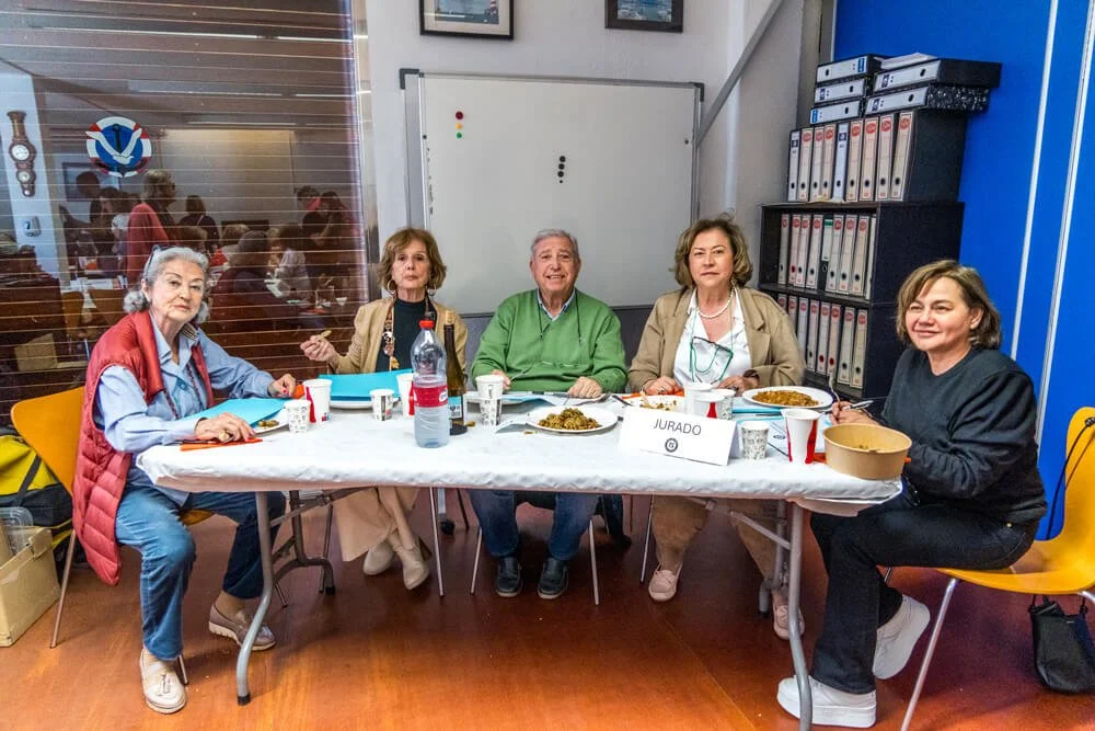 Grupo de adultos mayores sentados en una mesa con comida, en una sala con archivos y una pizarra, parecen estar en una reunión o evento social.