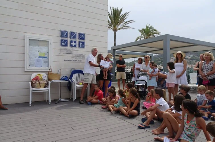 Grupo de niños y adultos en una ceremonia al aire libre en un área con sillones y palmeras, con una pared de fondo con signos de advertencia y un cartel de horario.