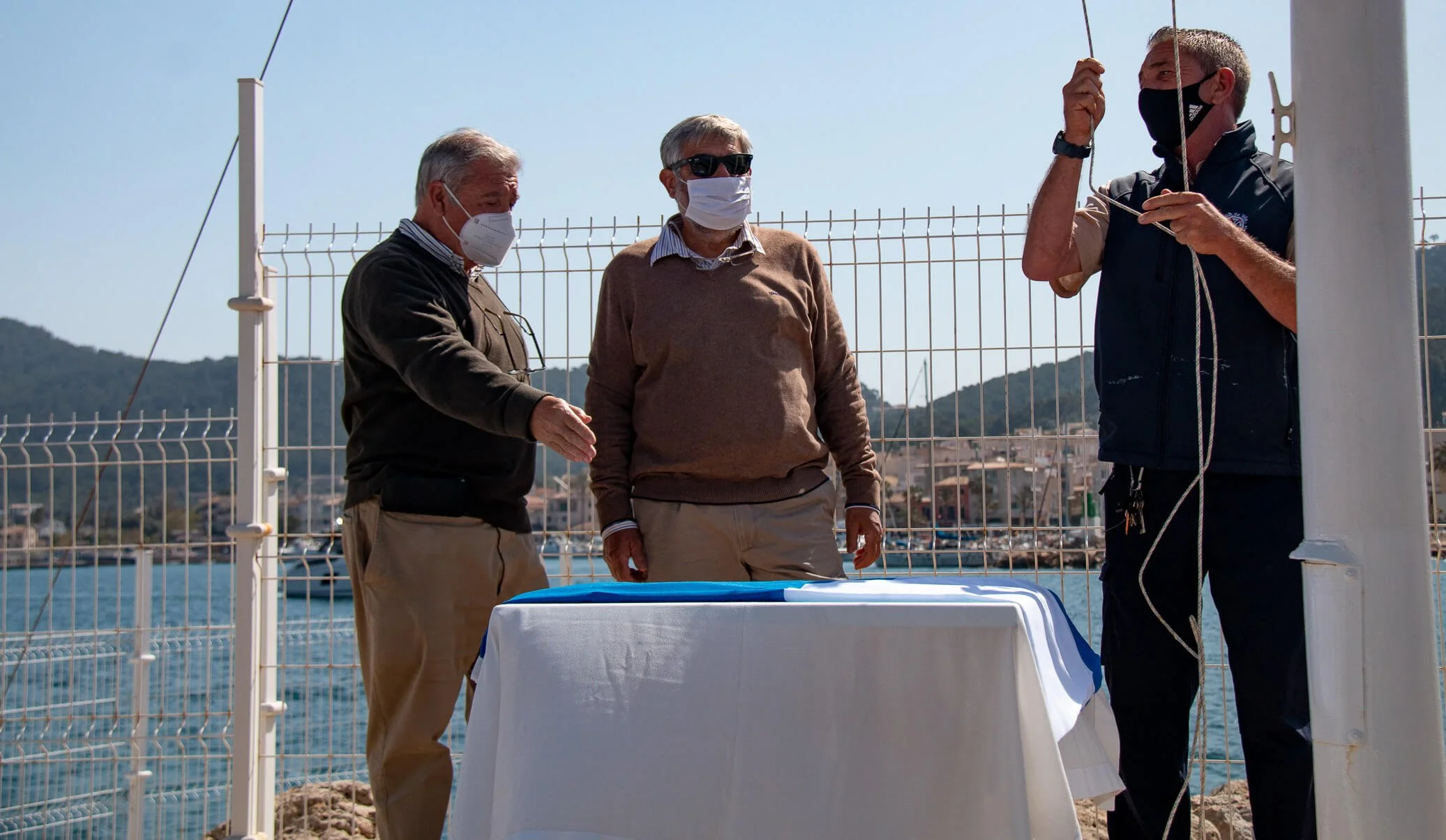 Tres hombres con mascarillas en una reunión al aire libre junto a un lago, con una mesa y una cerca de fondo.