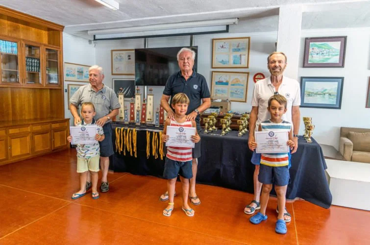Tres niños con certificados y tres adultos en un salón, con premios y diplomas en una mesa.
