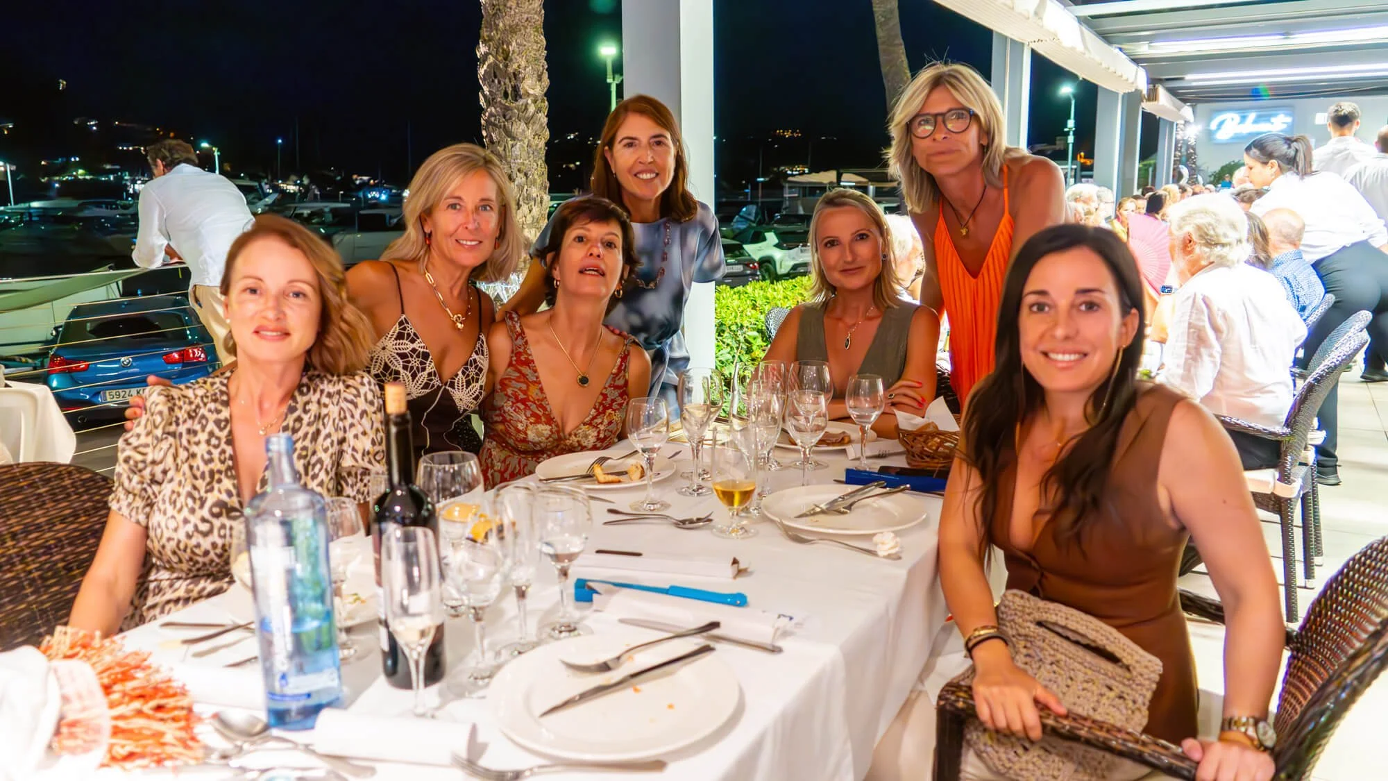 Grupo de diez mujeres sentadas y de pie en una mesa de restaurante al aire libre durante la noche, disfrutando de una comida y bebidas, con una decoración elegante y ambiente festivo.
