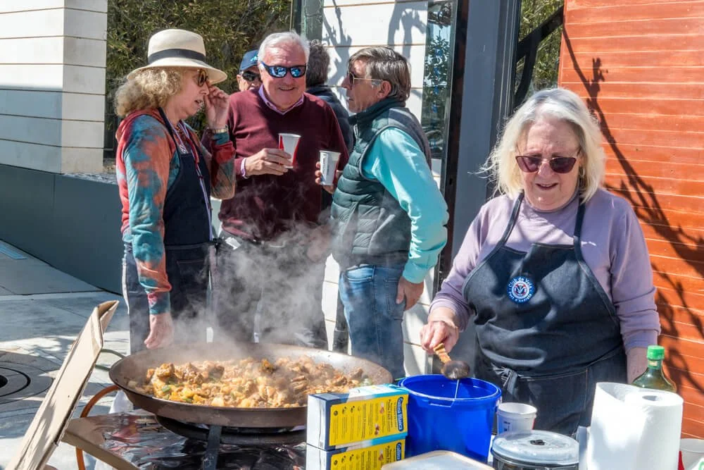 Un grupo de personas sonrientes disfrutando de una comida al aire libre, con una señora cocinando en una sartén grande sobre una mesa, rodeada de utensilios y otros adultos compartiendo y bebiendo.