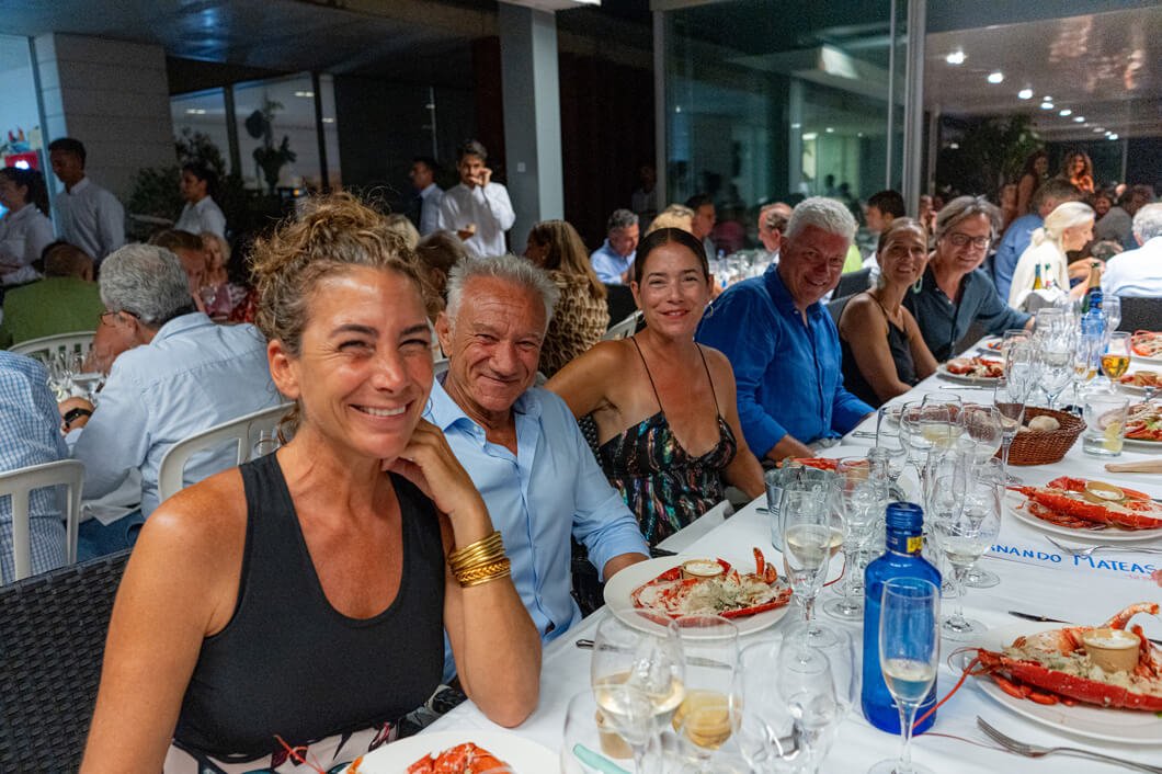 Personas sonriendo en una comida en un restaurante elegante, con platos de mariscos en una mesa larga y muchas personas en el fondo.