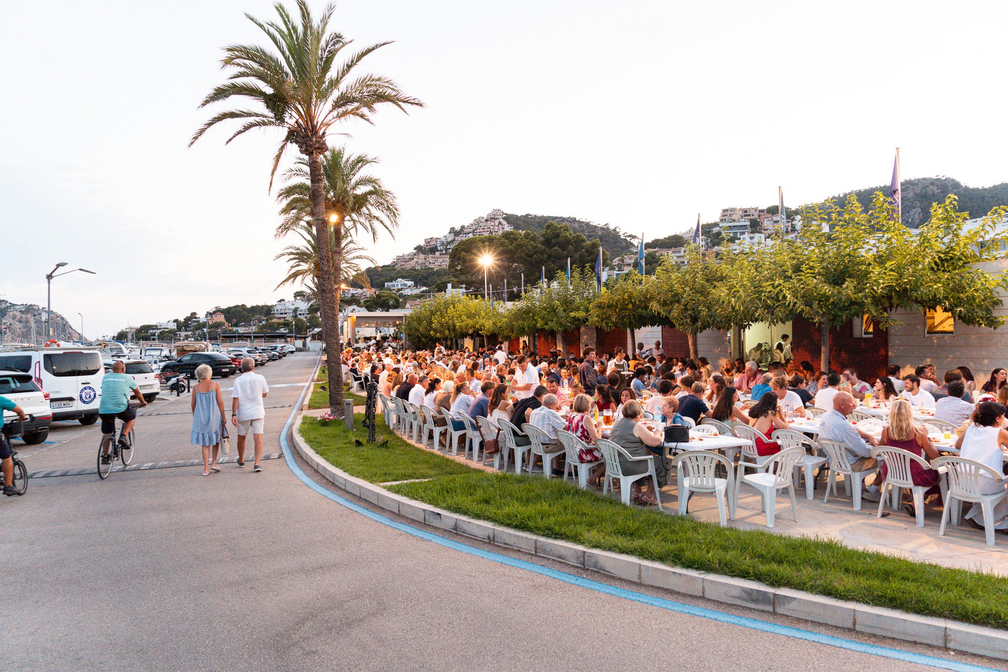 Una reunión o evento al aire libre con muchas personas sentadas en mesas, en un entorno costero con palmeras y colinas con casas en el fondo, durante el atardecer.