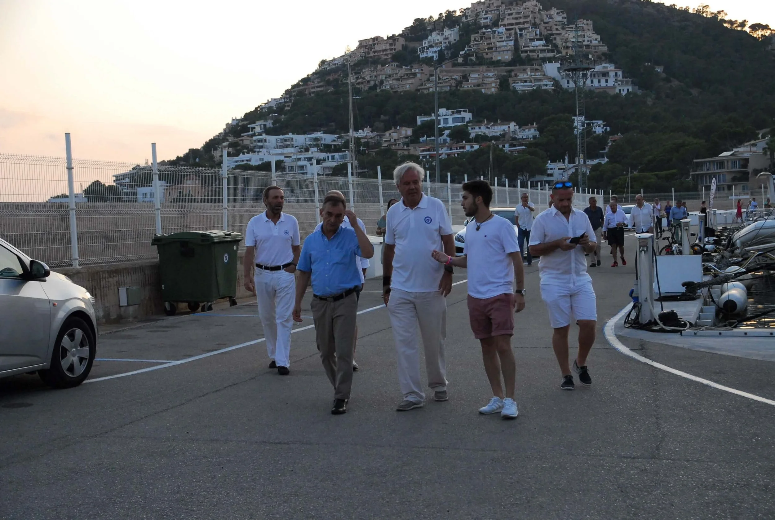 Grupo de personas caminando en un estacionamiento con colinas y casas en el fondo al atardecer.