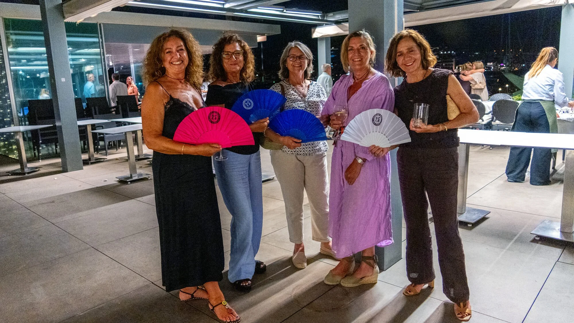 Cinco mujeres en una reunión social al aire libre en la noche, sosteniendo abanicos de mano de colores y bebidas, con fondo de ciudad y otras personas en el evento.