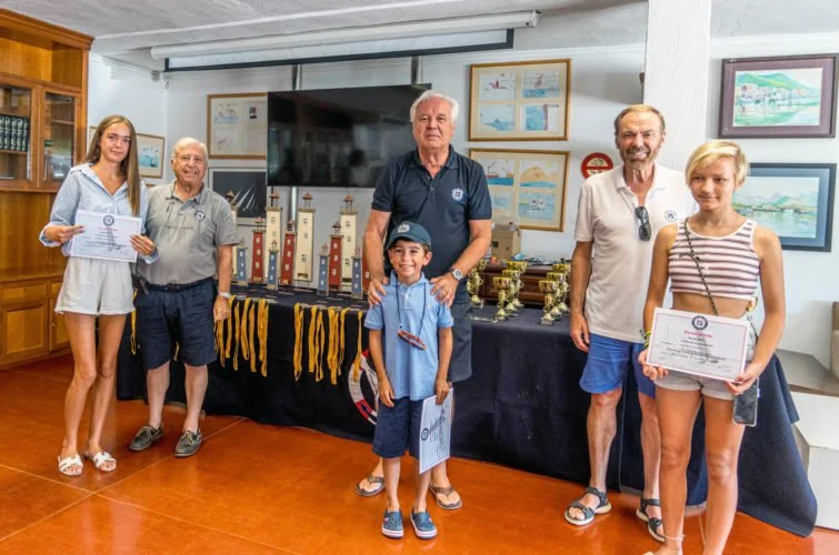 Grupo de personas en una ceremonia de premiación, con trofeos y certificados en una sala decorada con cuadros y modelos de faros en el fondo.
