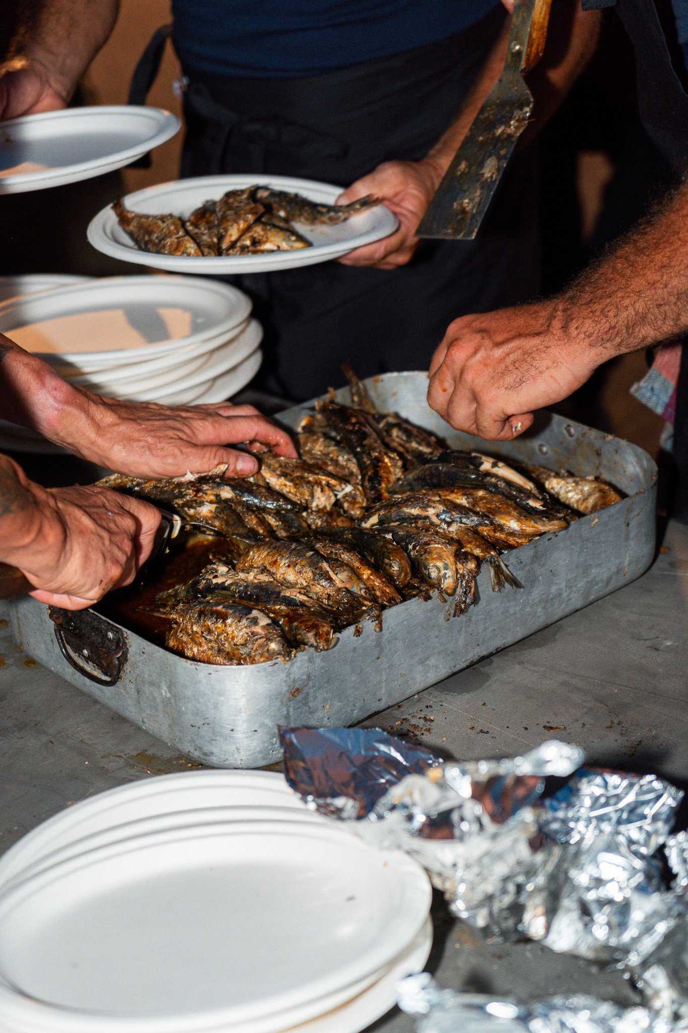Varias personas sirviendo pescado asado en platos blancos, mesas con más platos y papel aluminio en un ambiente de comida compartida.