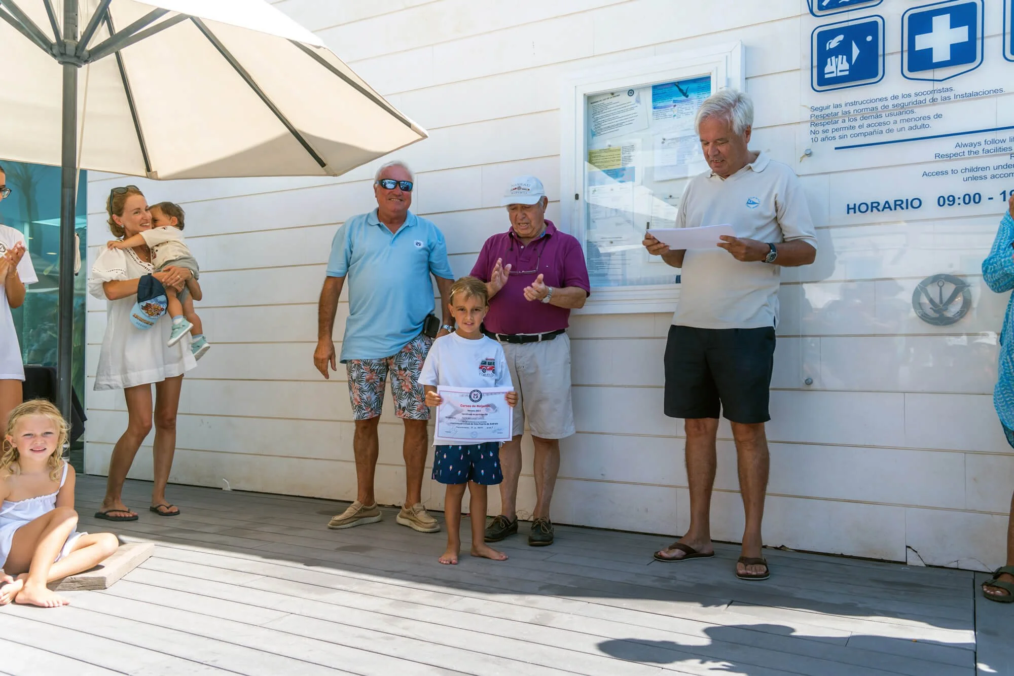 Grupo de personas en una ceremonia al aire libre, con niños y adultos, algunas sosteniendo certificados, en un lugar con paredes blancas y cartelera de instrucciones de seguridad y horarios.
