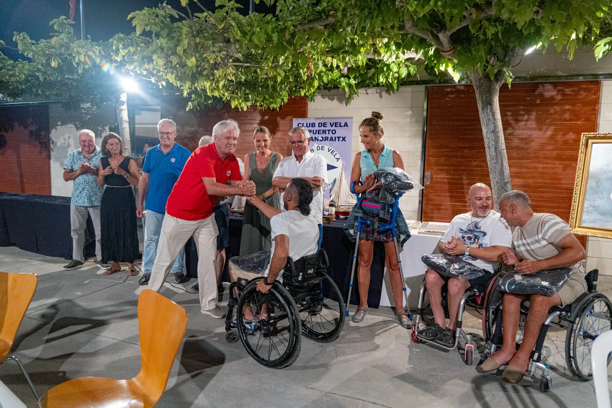 Personas en una reunión nocturna, algunas en sillas de ruedas, intercambiando un apretón de manos y sonriendo, en un espacio abierto con árboles y decoración.