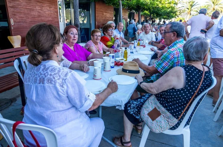 Grupo de personas mayores sentadas en una mesa al aire libre, disfrutando de una comida en una reunión social.