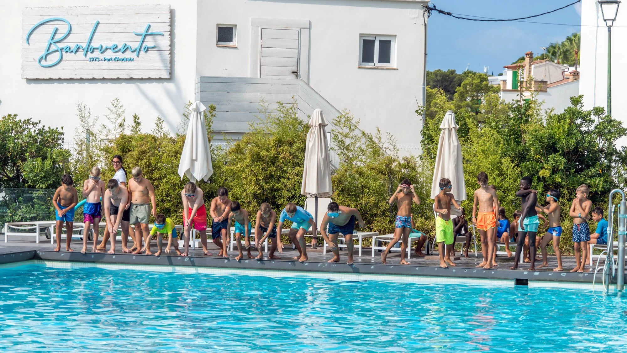 Niños y niñas con trajes de baño en la piscina, algunos en la orilla y otros en el agua, con un adulto en el fondo y vegetación detrás, en una zona de recreo acuático en un día soleado.