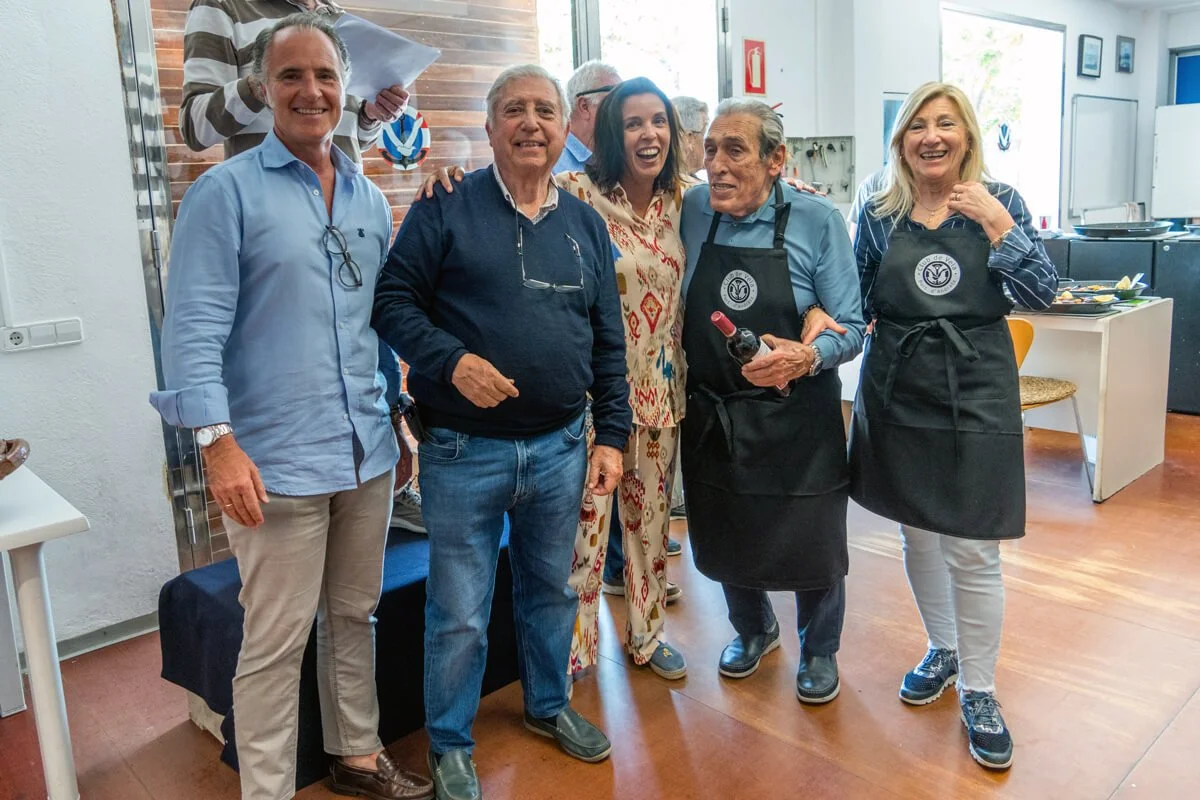 Grupo de seis personas sonriendo en una cocina, con dos de ellas usando delantales, una de ellas sosteniendo una botella de vino.
