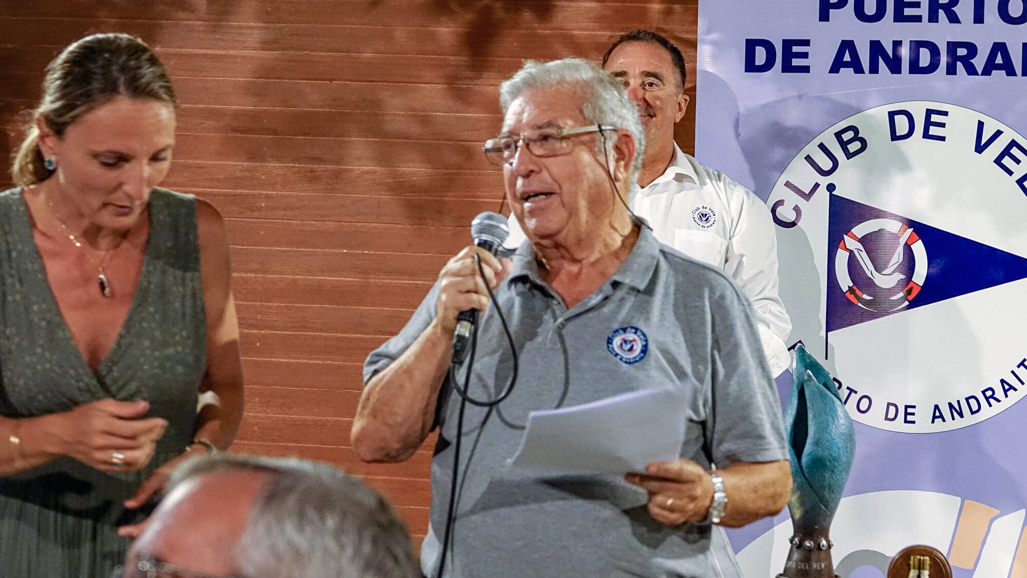 Un grupo de personas en una reunión o evento, con un cartel que dice 'Club de Veleros de Andratx' en el fondo. Una persona mayor con gafas y camiseta de manga larga está hablando con un micrófono, mientras que dos personas lo escuchan y parecen parti