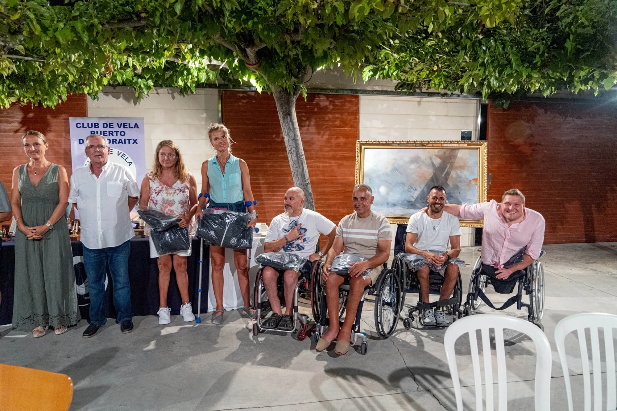 Personas en sillas de ruedas y otras de pie, durante una ceremonia o evento en una zona cubierta, con un cartel que dice 'Club de vela Puerto D'Airaix', un árbol grande y un cuadro grande en la pared de fondo.