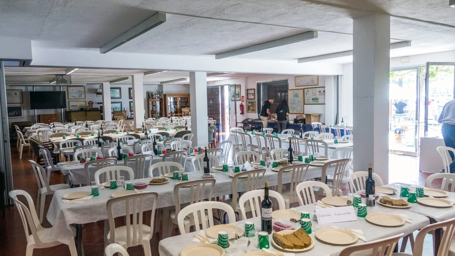 Sala de fiestas preparada con mesas y sillas blancas, decoradas con platos, cubiertos, botellas de vino, galletas y vasos verdes, lista para un evento o comida.