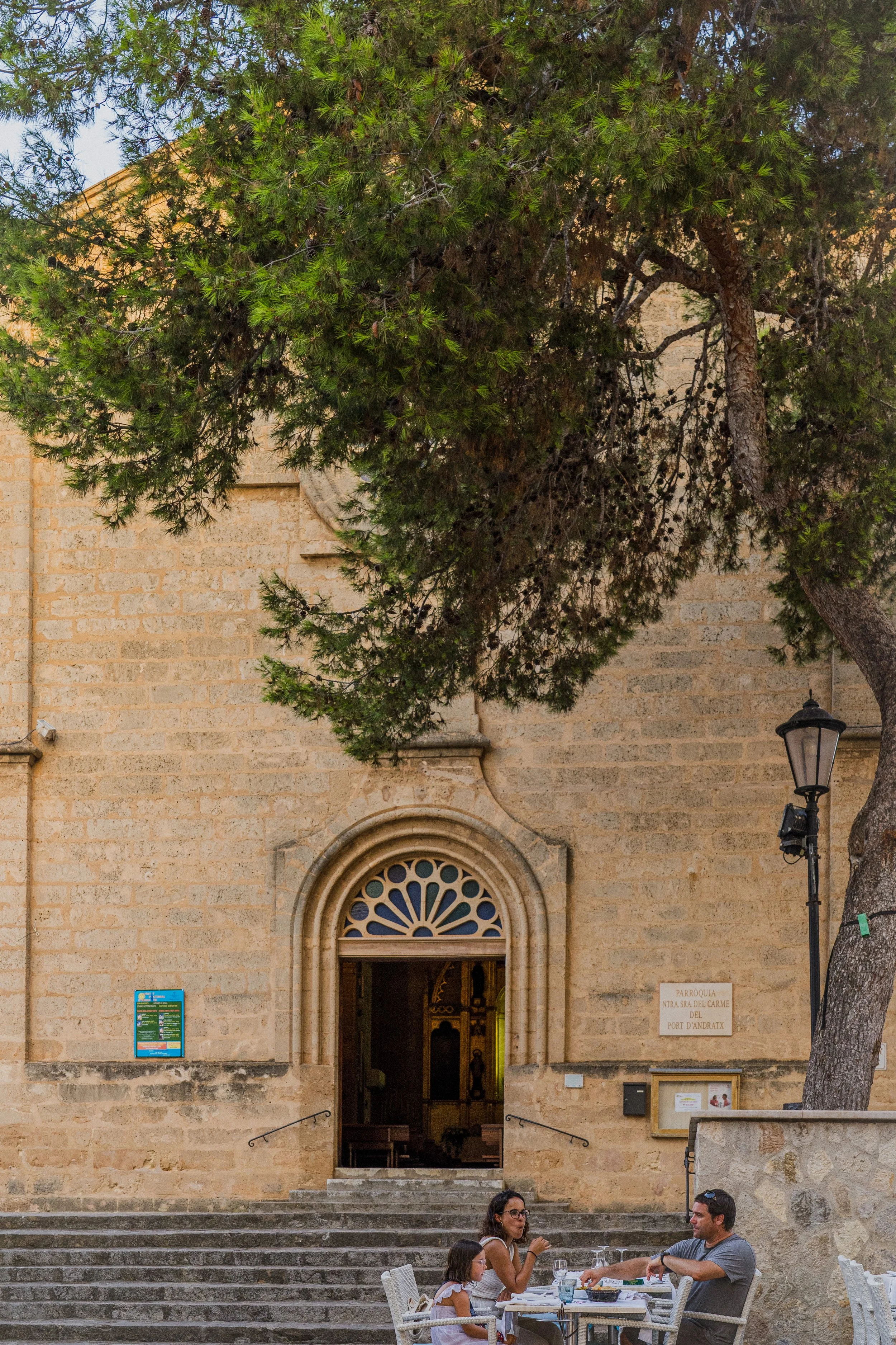 Grupo de personas sentadas en una terraza frente a una iglesia antigua de ladrillo, con un árbol grande y una farola en primer plano, en una ciudad histórica.