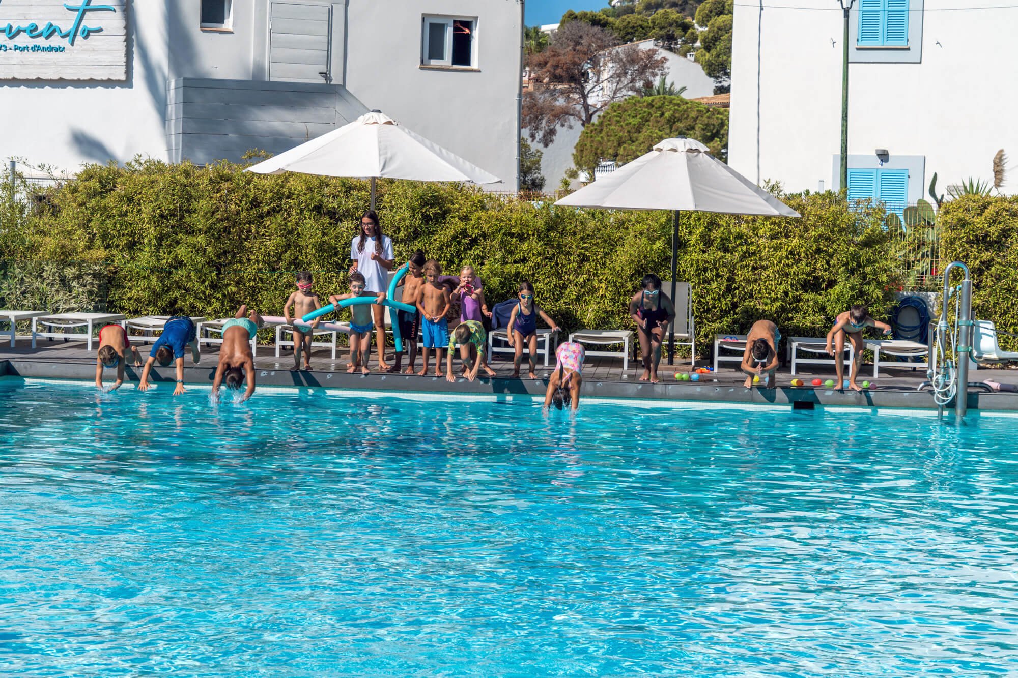 Niños y jóvenes en la piscina, algunos saltando al agua, otros en la orilla, supervisados por una mujer. Hay sombrillas blancas y vegetación de fondo.