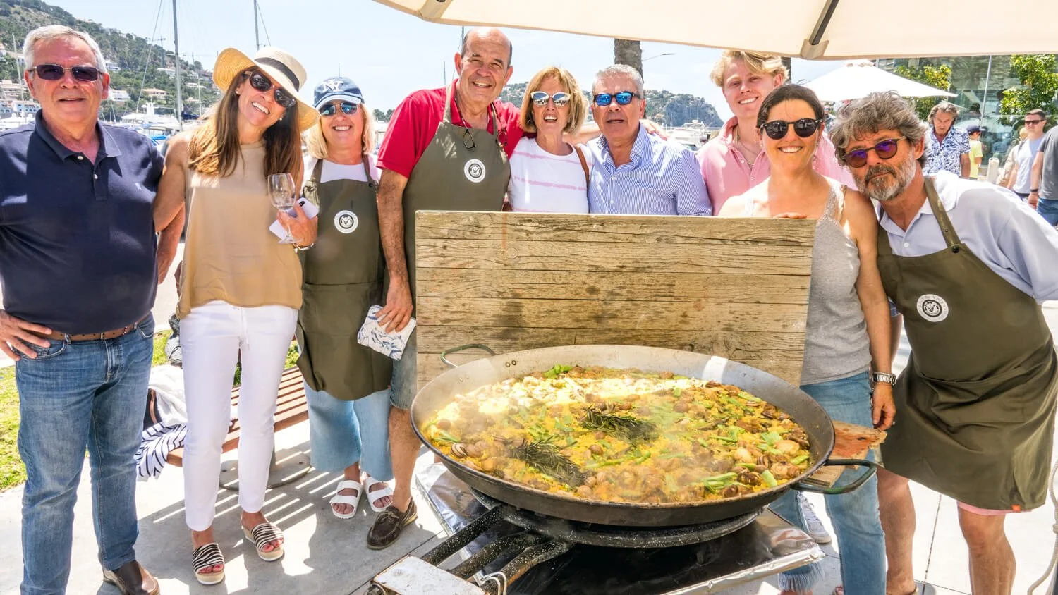 Grupo de personas sonriendo en una celebración al aire libre con una gran paella en una sartén gigante en primer plano.