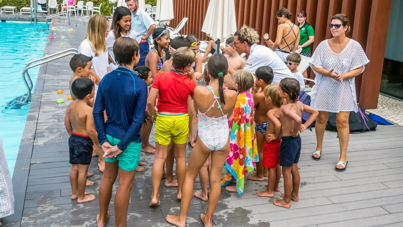 Grupo de niños y adultos en la zona de la piscina, algunos con trajes de baño, participando en actividades y juegos acuáticos, en un ambiente de recreación y diversión cerca de la piscina.