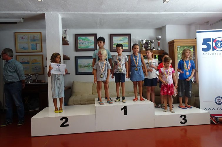 Niños en un podio de premiación, con medallas, en una ceremonia en interiores. Hay una mujer mayor con gafas a la izquierda y un banner en el lado derecho que conmemora el 50 aniversario.