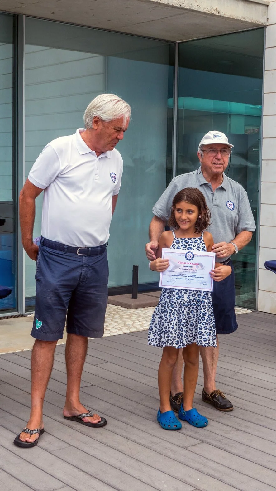 Una niña recibe un certificado de natación de dos hombres adultos, uno con camisa blanca y pantalones cortos, y otro con sombrero y camisa gris, en un entorno exterior con ventanas de vidrio.