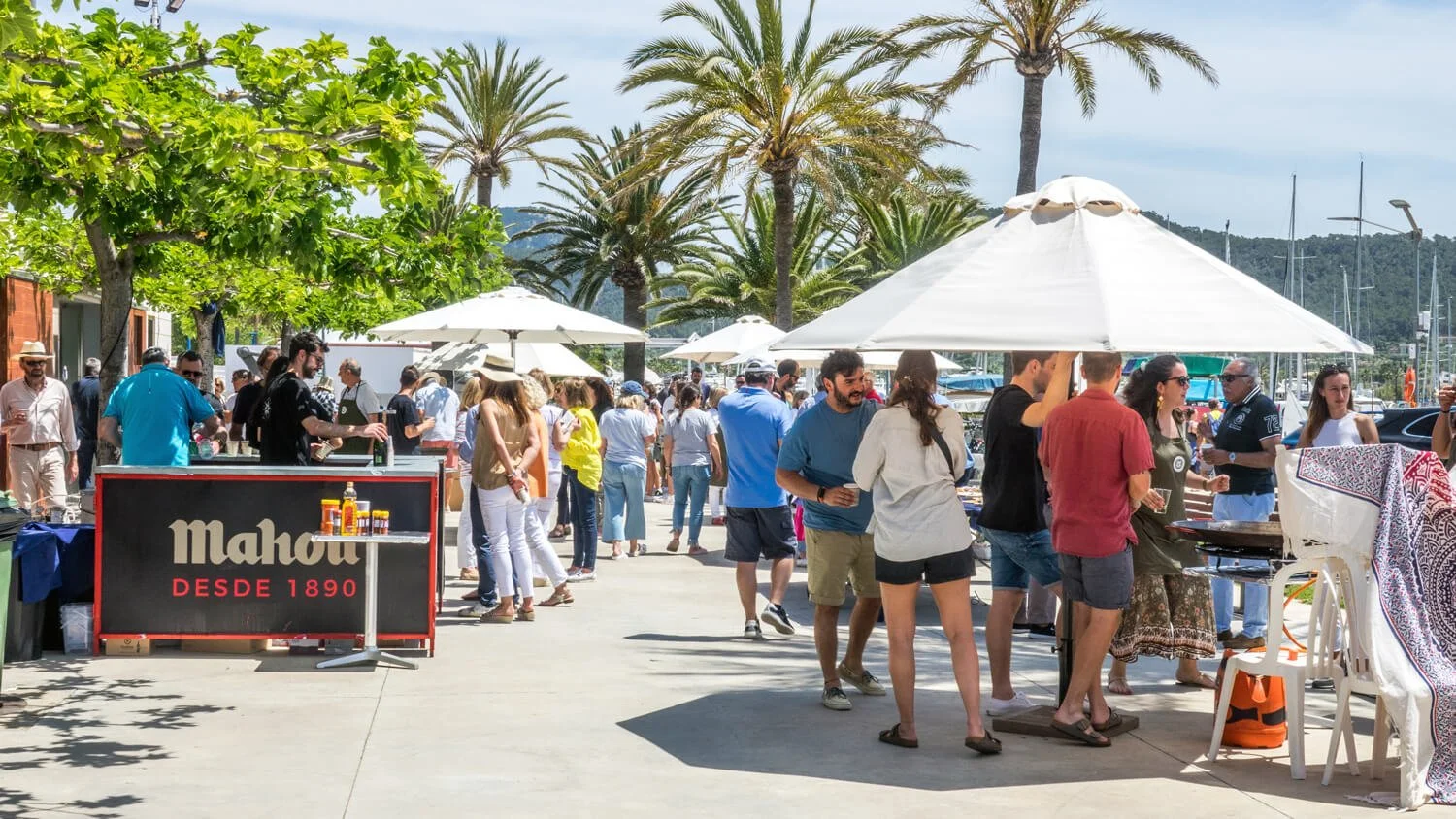 Personas en una feria o mercado en un lugar con árboles y palmeras, algunos con sombrillas, vendiendo y comprando productos en un día soleado junto a una marina.