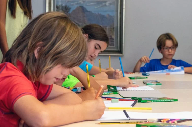 Niños en una mesa, dibujando con lápices de colores, en un entorno de aula o taller.